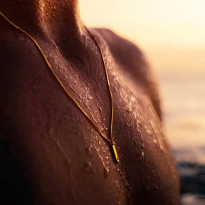 Homem usando colar dourado fino com pingente geométrico retangular na praia.