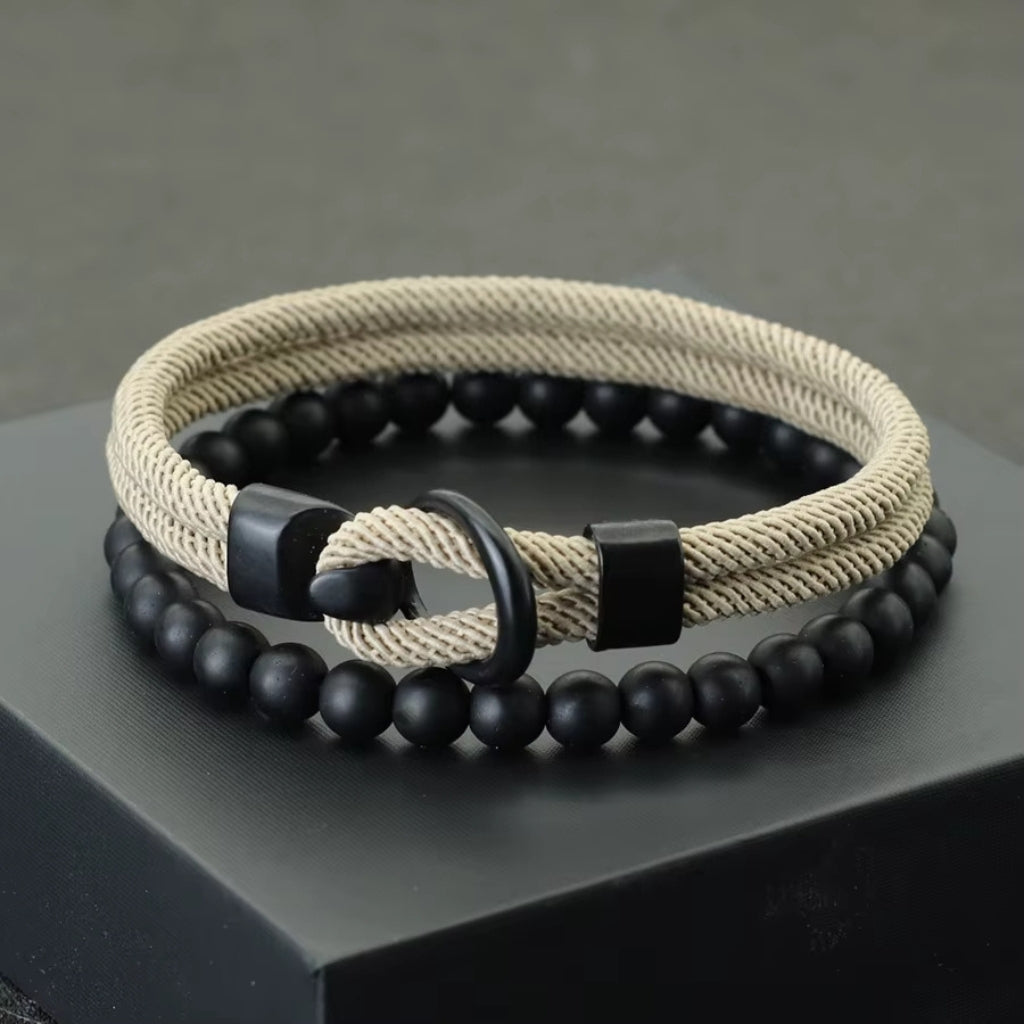Kit de pulseiras masculinas Milano: uma de corda trançada bege e outra de pedras pretas foscas sobre uma caixa preta.